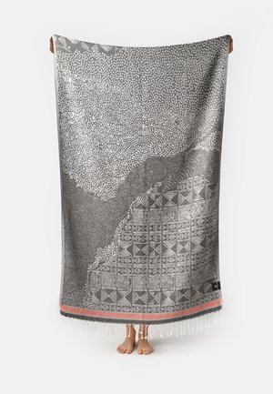 Person hält ein großes grau-weiß gemustertes Textil mit geometrischen Designs und fransigen Rändern, barfuß mit sichtbaren Füßen darunter.