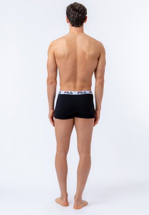 Man staat blootsvoets, met de rug naar de camera, draagt zwarte boxershorts met een witte FILA-logo tailleband tegen een effen witte achtergrond.