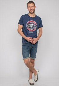 T-shirt en coton bleu marine avec un motif de moto, associé à un short en denim, avec des manches retroussées et des chaussures à enfiler de couleur claire.