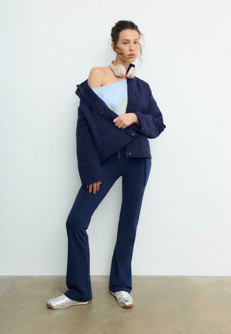 Jeune femme portant une veste bleu marine tombée sur une épaule, un haut bleu clair, un pantalon bleu marine, des baskets argentées et un casque beige autour du cou, debout contre un mur blanc.