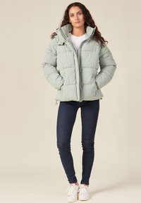Giacca puffer verde menta con design trapuntato, chiusure a zip e bottoni a pressione, cappuccio regolabile e orlo elastico. Indossata con jeans scuri e sneakers bianche.