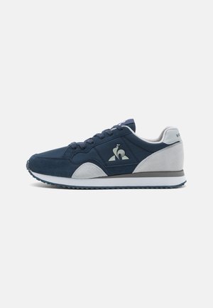 Scarpe sportive navy e grigie con materiali in suede e sintetici, dotate di punta arrotondata, chiusura con lacci e un accento logo sul lato.