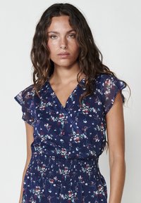 Rochie florală marină cu mâneci scurte tip fluture, bust ajustat și talie plisată. Prezintă un model floral multicolor pe întreaga suprafață. Țesătură ușoară.