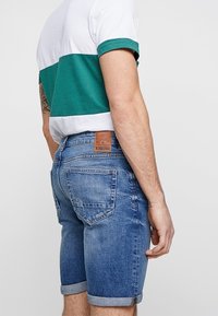 Sinisiä farkkushortseja, joissa on käärityt lahkeet, ruskea nahkapaikka, takataskut ja haalistunut pesu. Yhdistetty vihreän ja valkoisen raidallisen paidan kanssa.