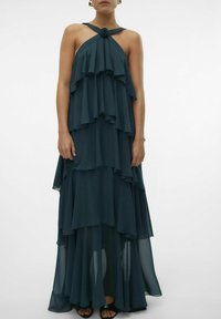 Tirkizna maxi haljina od laganog chiffon materijala s halter izrezom i ružinim akcentom, s lepršavim volanima i opuštenim krojem.