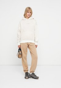 Hanorac dungat crem cu linii bej, pantaloni de trening bej și pantofi sport gri. Ține în mână o geantă mică neagră și bej. Ținută casual.