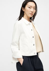 Γυναίκα με ίσια σκουρόχρωμα μαλλιά φοράει λευκό σακάκι με κουμπιά πάνω από μπεζ μπλουζάκι και μαύρο παντελόνι, πόζαρε με το ένα χέρι στην τσέπη.