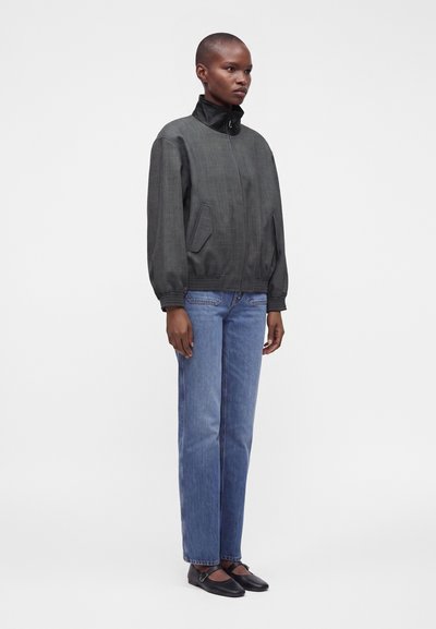 Femme aux cheveux courts portant une veste bomber gris foncé, un jean bleu droit et des chaussures Mary Jane noires, debout devant un fond clair.