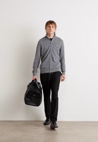 Gray zip-up sweater, black pants, and shoes. Carrying a black leather duffel bag. Simple, modern design against a neutral background.