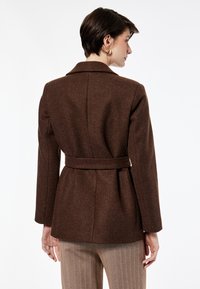 Chaqueta marrón de mezcla de lana con cintura ceñida con cinturón, cuello con muesca y mangas largas. Textura suave, corte entallado y detalle de abertura trasera.