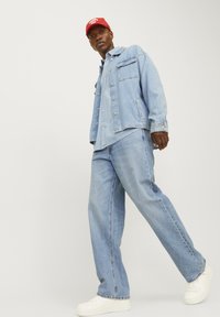 Lichtblauwe denimjack met twee borstzakken, gecombineerd met bijpassende lichtblauwe jeans en witte sneakers. Een rode pet wordt op het hoofd gedragen.