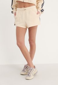 Shorts côtelés de couleur crème avec taille élastique, poches latérales et bandes latérales sombres contrastantes. Associés à des baskets de sport claires.