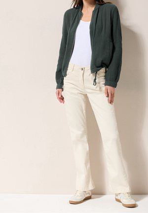 Femme portant une veste zippée vert foncé, un débardeur blanc, un pantalon couleur crème et des baskets blanches, debout contre un mur beige.