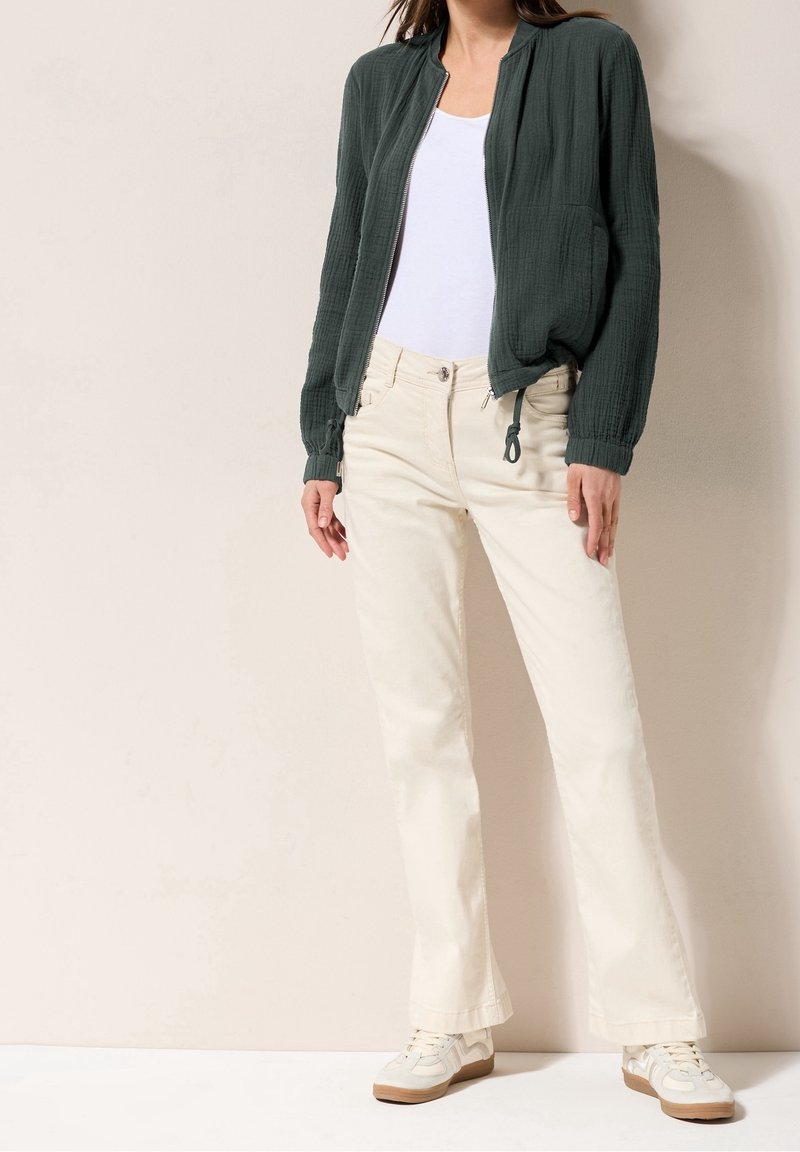 Femme portant une veste zippée vert foncé, un débardeur blanc, un pantalon couleur crème et des baskets blanches, debout contre un mur beige.