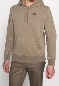 Felpa beige con cappuccio, tasca a marsupio, tessuto testurizzato, polsini a costine e un piccolo logo sul petto. Morbida e con una vestibilità rilassata.