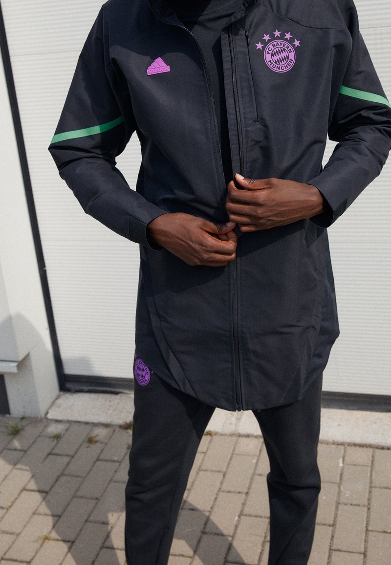 Personne fermant la fermeture éclair d'une veste noire du FC Bayern Munich avec un logo violet et des bandes vertes, debout sur un sol pavé en extérieur.