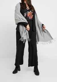 Sciarpa di lana grigia con bordi frangiati drappeggiata su una blusa nera con ricami floreali, abbinata a pantaloni neri a gamba larga e stivali con tacco.