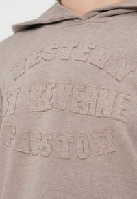 Bež pulover s kapuco z izbočenimi všitimi črkami, ki na prsih kažejo napis "Western 87 Kverne Paiston".