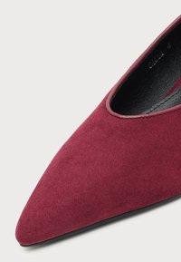 Chaussure plate à bout pointu recouverte de suède bordeaux avec un bord lisse autour de l’ouverture de la chaussure et une doublure intérieure noire.