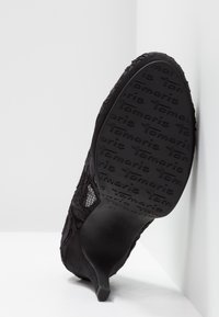 Chaussure à talon haut noire inclinée vers le haut avec une semelle texturée arborant la marque répétée « Tamaris » et des détails en dentelle et mesh sur la tige.