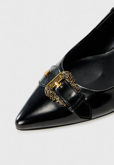 Chaussure plate en cuir noir avec bout pointu, présentant une boucle ornée en or et un détail de sangle sur le dessus. Texture lisse et brillante.