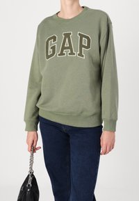 Světle olivový svetr s kulatým výstřihem, žebrovanými manžetami a velkým logem "GAP" v strukturovaném bílém písmu. Spárováno s tmavě modrými džíny a černou taškou.