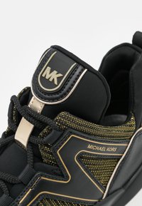 Schwarze Sneaker mit goldenen Akzenten, einem strukturierten Obermaterial aus schwarzen und goldenen Fäden sowie einem Logo auf der Zunge. Mit Schnürsenkeln verschlossen.