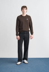 Ruskea collegepaita, jossa on resorit hihansuissa ja pyöreä pääntie, yhdistettynä leveälahkeisiin mustiin farkkuihin ja valkoisiin lenkkareihin, seisomassa sinisellä matolla.