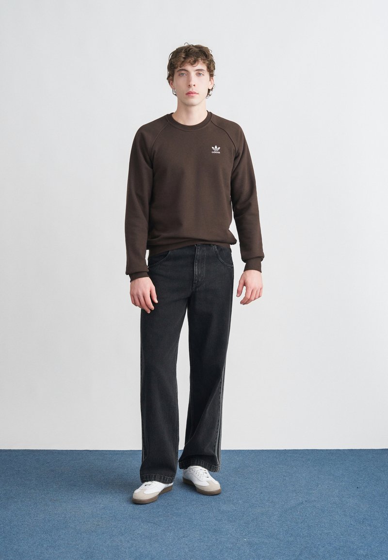 Brun sweatshirt med ribbade muddar och rund halsringning, kombinerad med vida svarta jeans och vita sneakers, stående på blå matta.