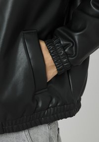 Veste en similicuir noir avec un col et des poignets côtelés, dotée d'une poche latérale et d'un ourlet froncé pour un look ajusté.