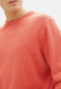 Koralfarvet strikket sweater med rund hals og ribbede manchetter, prydet med et diskret logo på brystet og en glat tekstur.