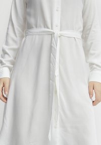 Robe chemise blanche à manches longues, fabriquée dans un tissu léger, avec un devant boutonné, une ceinture à nouer à la taille, et une texture lisse.