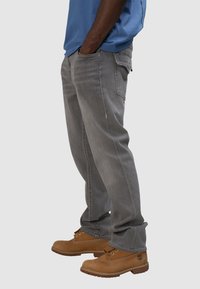 Vaqueros de mezclilla gris con un corte relajado y cinco bolsillos, combinados con botas de trabajo marrones. Los pantalones presentan costuras visibles y un ligero desgaste.