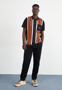 Camisa de manga corta con rayas verticales en naranja, blanco y negro, que presenta un bolsillo en el pecho. Combinada con pantalones de pana negros y zapatillas beige.
