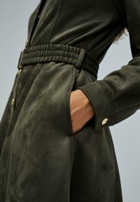 Robe en daim vert olive avec ceinture, devant boutonné, poches latérales et tissu texturé, présentant des boutons dorés aux poignets.