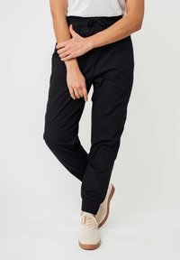 Schwarze Jogginghosen mit elastischem Bund und Kordelzug, schmal zulaufende Beine und gerippte Bündchen. Getragen mit hellen Turnschuhen.