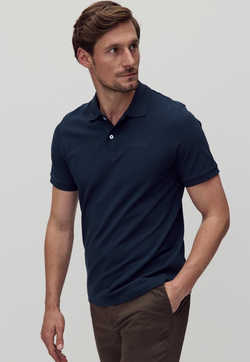 Mörkblå pikétröja med kort ärm och krage, två knappslå, samt en subtil logotyp broderad på bröstet. Bruna byxor.