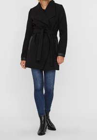 Schwarzer Stoffmantel mit tailliertem Gürtel, großem Kragen und Lederakzenten an den Ärmeln. Kombiniert mit blauen Jeans und schwarzen Stiefeletten.