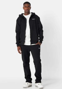 Sudadera con cremallera negra y pantalones de chándal a juego, fabricados con tejido suave. La sudadera cuenta con bolsillos frontales, mientras que los pantalones tienen un pequeño logo en el muslo.