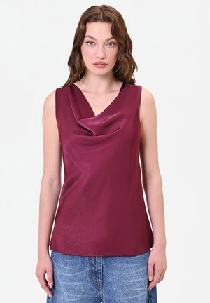 Jeune femme aux cheveux bruns ondulés porte un haut bordeaux brillant sans manches et une jupe en denim bleue sur un fond clair uni.