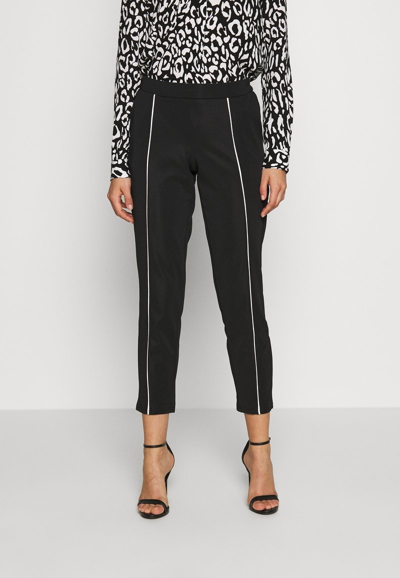 Femme portant un pantalon court noir à rayures blanches verticales et des talons noirs à bride à la cheville, assorti d’un haut à manches longues à motif noir et blanc.