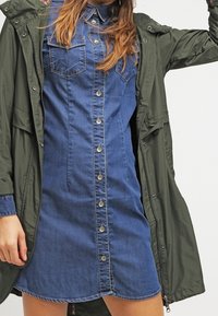 Denim-mekko, jossa on napitettavat etuosat, terävä kaulus ja rintataskut, kerroksittain oliivinvihreän työjakun alla.