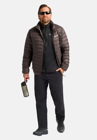 Puffer jacket in dark brown with horizontal stitching, black inner layer, black pants, and rugged black shoes. Holding a green water bottle.