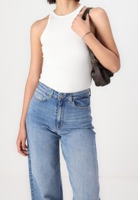 Person wearing sleeveless white top and blue jeans, holding a dark brown shoulder bag, with a faint tattoo on their wrist.