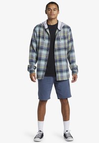 Karierte Flanellhemd in grün und blauen Tönen, mit einer grauen Kapuze. Kombiniert mit blauen Shorts und schwarzen Sneakers, ergänzt durch weiße Socken.