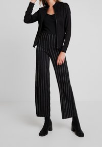 Giacca nera con zip sopra una canotta nera, abbinata a pantaloni a vita alta e a gamba larga a righe con strisce argentate e stivali neri con tacco.