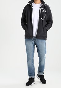 Henkilö, joka on pukeutunut mustaan Nike-tuulitakkiin, valkoiseen t-paitaan, vaaleansinisiin farkkuihin, joissa on lahkeet, ja mustiin lenkkareihin, seisoo kädet taskuissa.