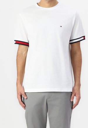 Mies, jolla on valkoinen lyhythihainen t-paita, jossa on punaiset, valkoiset ja laivastonsiniset raidalliset hihansuut ja pieni rintalogo, yhdistettynä harmaisiin housuihin.