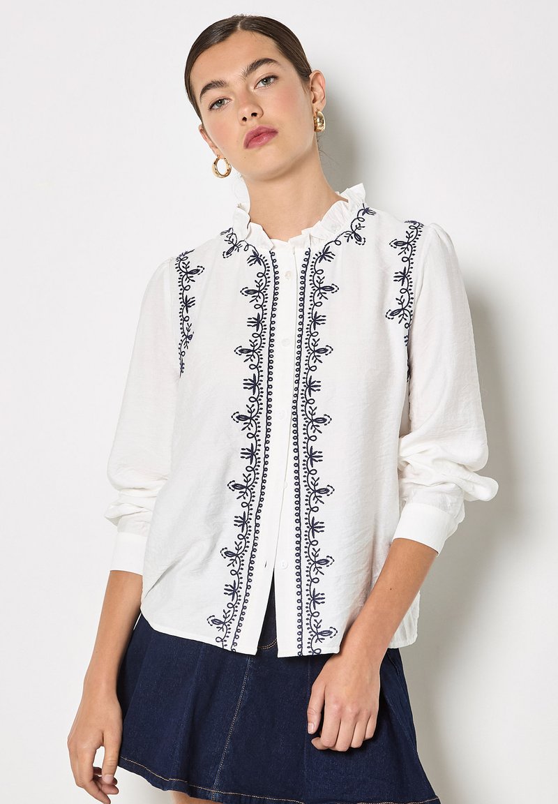 Jeune femme portant un chemisier blanc avec une broderie noire et un col à volants, associé à une jupe en denim foncé, debout contre un mur blanc.