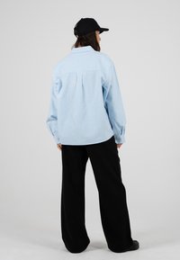 Femme portant une casquette de baseball noire, une chemise ample rayée bleu clair et un pantalon noir large, debout face à un fond gris.